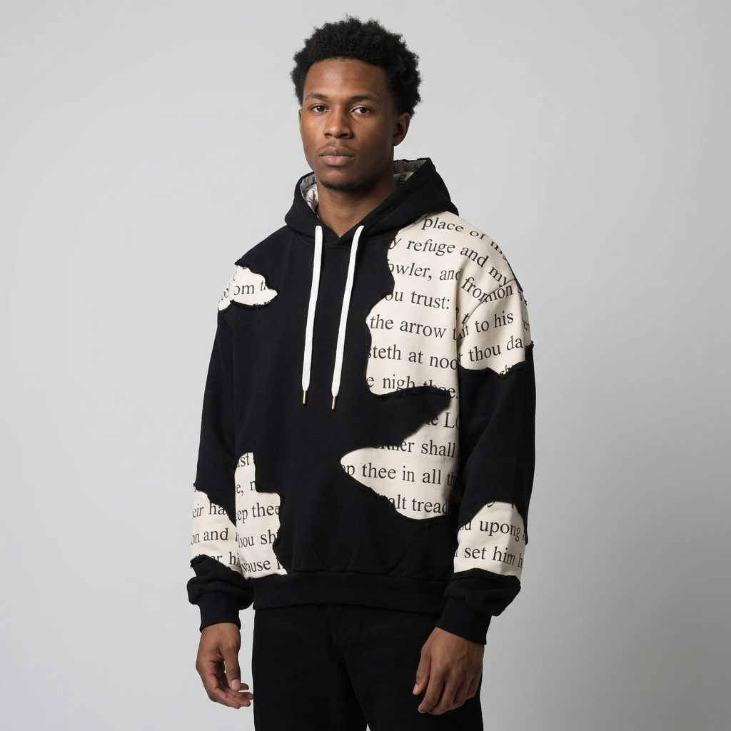 A man wearing a black pullover hoodie with large beige patches of printed text, posed against a neutral light gray background.